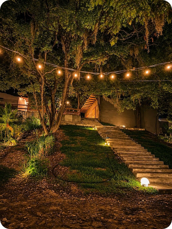 Un espacio exterior nocturno con una escalera de piedra que conduce a una estructura de madera, rodeado de árboles y luces colgantes.