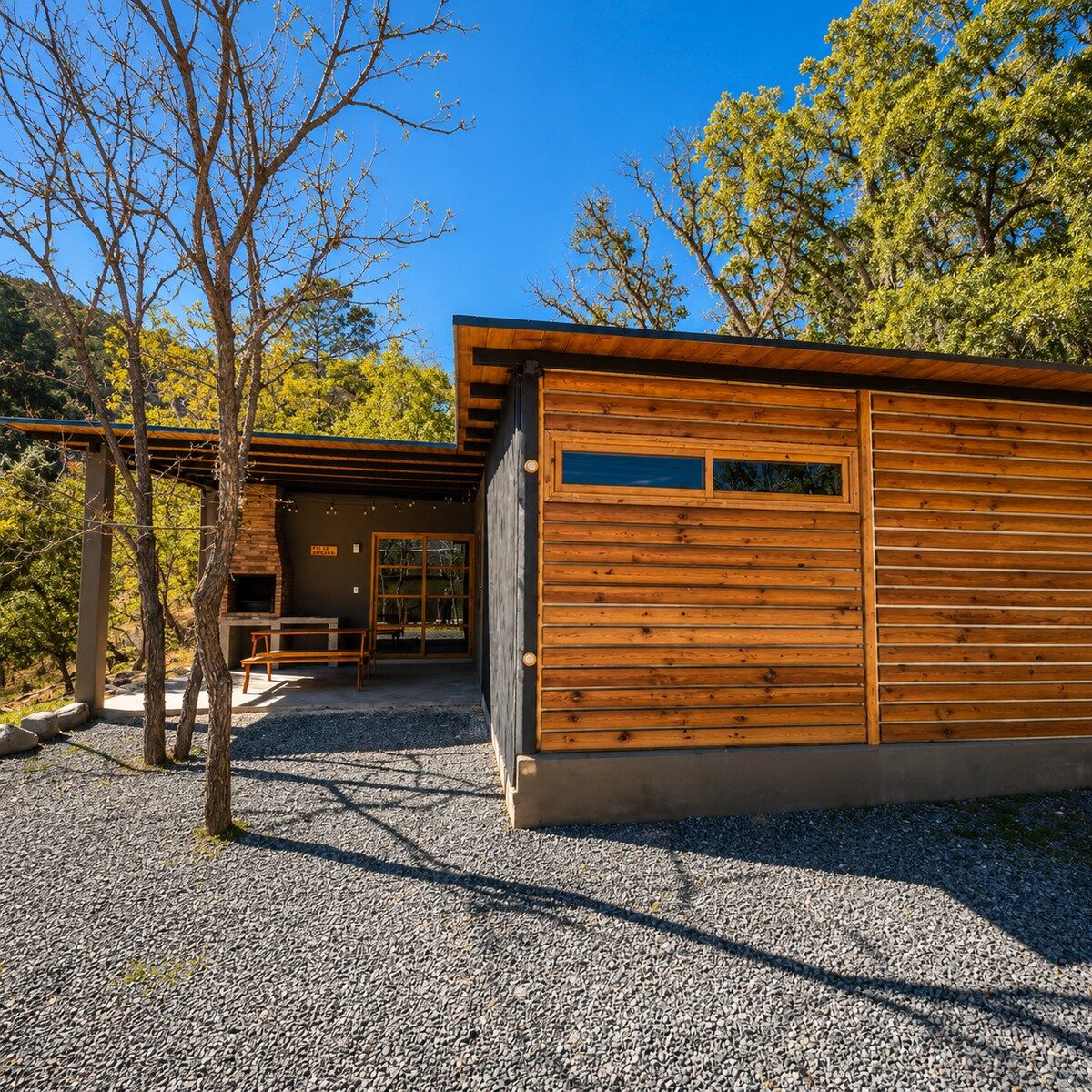 A modern wooden cabin with a covered patio, a stone fireplace, and a gravel driveway.