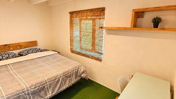 A room with a bed, a small table, a window with blinds, and a shelf with a plant.