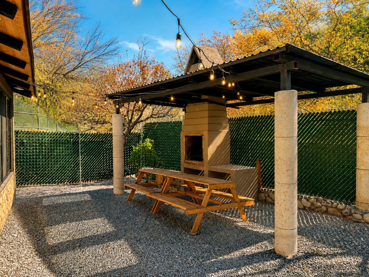 A covered outdoor seating area with a grill, wooden picnic table, and string lights.