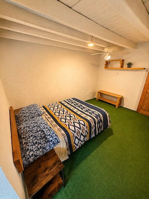 A small, cozy room with a single bed, a wooden bench, and a ceiling fan.
