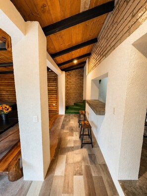 Ein Flur mit Holzboden, weißen Wänden und einer Backsteinwand. Zwei Hocker und ein Blumentopf sind ebenfalls zu sehen.