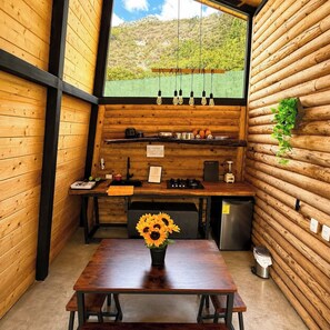 A rustic kitchen with wooden walls, a wooden table with a sunflower centerpiece, and a view of a green landscape through a window. - VILLA AMADA. Cabaña Alba (Santiago)