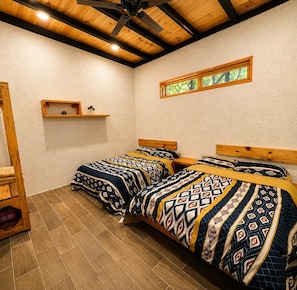 A room with two beds, wooden headboards, a shelf with a plant, and a window with a view of greenery.