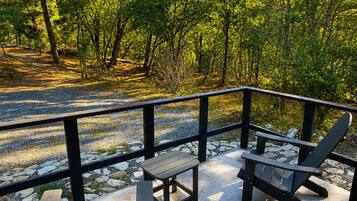 Área externa com assentos Adirondack e uma mesa, cercada por um deck de madeira e vista para uma área arborizada.