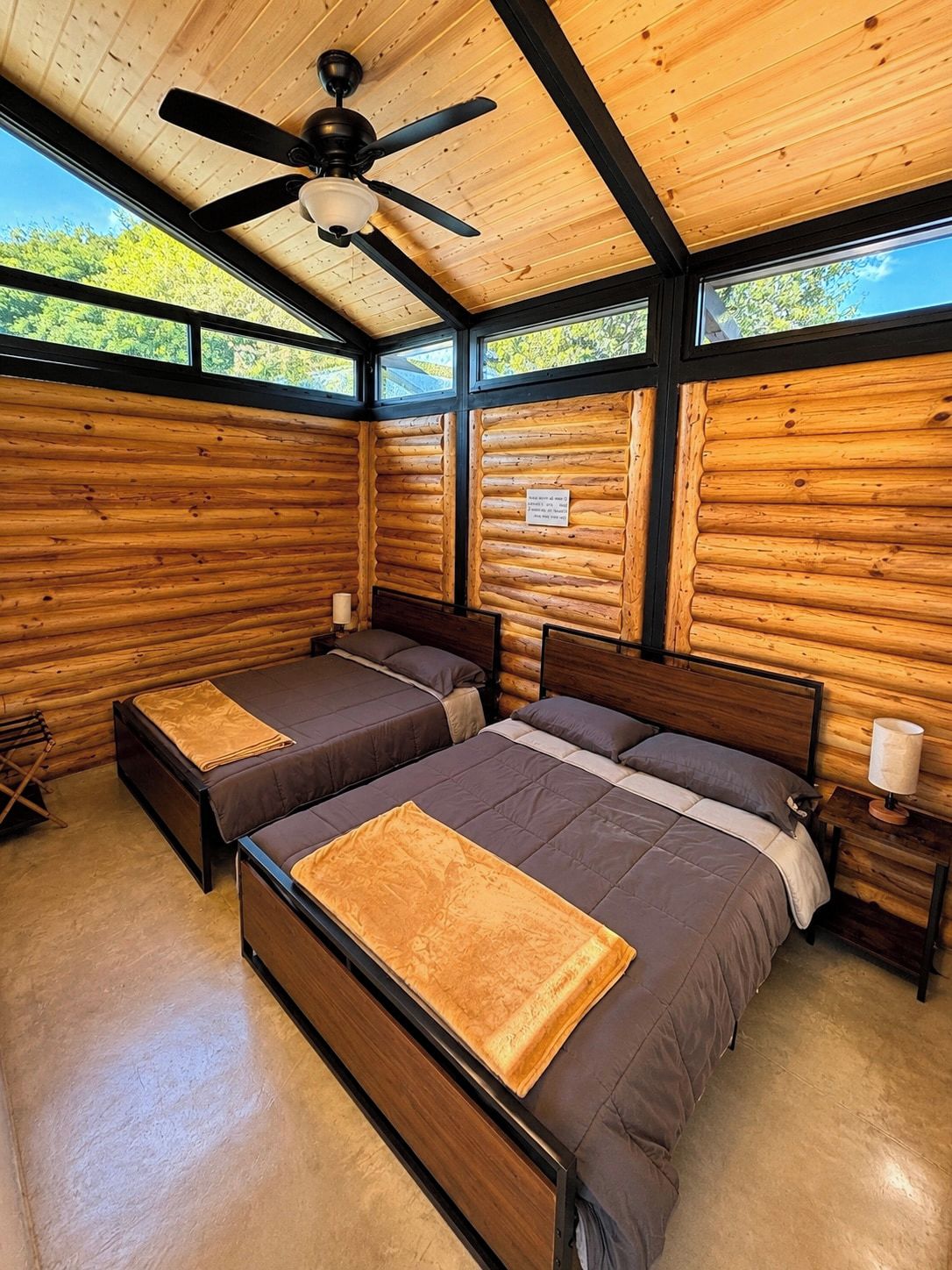 Ein Schlafzimmer in einer Blockhütte mit zwei Betten, einem Deckenventilator und einem Fenster mit Blick auf Bäume.