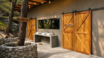 Un baño con dos puertas de madera que dicen 'BAÑOS' y 'MUJERES', un banco y una jardinera de piedra.