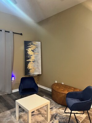 A modern living room with a white table, two blue chairs, a brown ottoman, and a large abstract painting on the wall. - Modern Gulfport Retreat. Stylish 1 BR, Full Kitchen, Patio & Private Driveway (Gulfport)