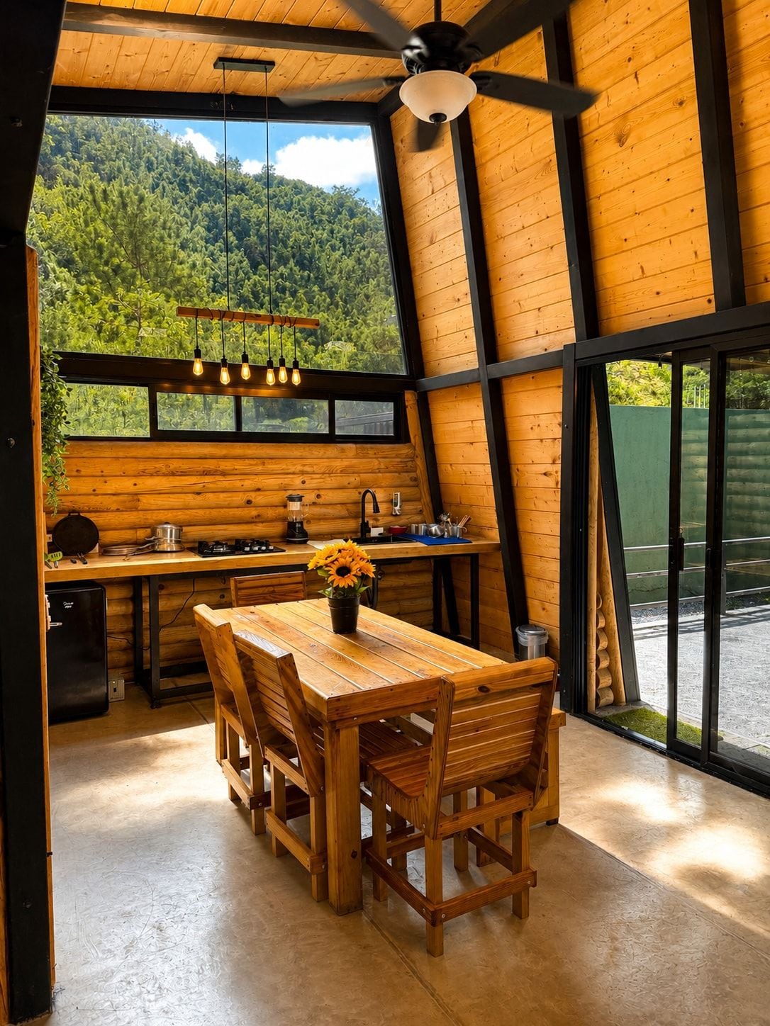 A wooden interior with a dining table, chairs, a ceiling fan, and a view of a forest through large windows.