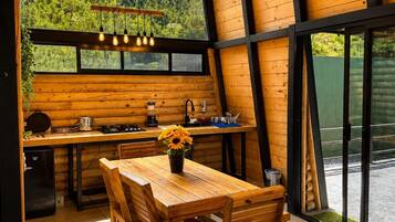 A wooden interior with a dining table, chairs, a ceiling fan, and a view of a forest through large windows.