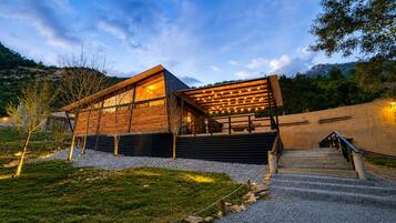 Un edificio moderno con fachada de madera, un área exterior techada con luces colgantes y un camino de grava que lleva a una escalera.