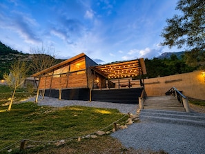 Un edificio moderno con fachada de madera, un área exterior techada con luces colgantes y un camino de grava que lleva a una escalera.