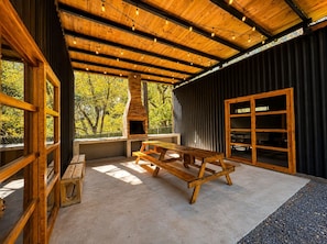 A rustic wooden interior with a large table and benches, a brick fireplace, and large windows offering a view of trees.