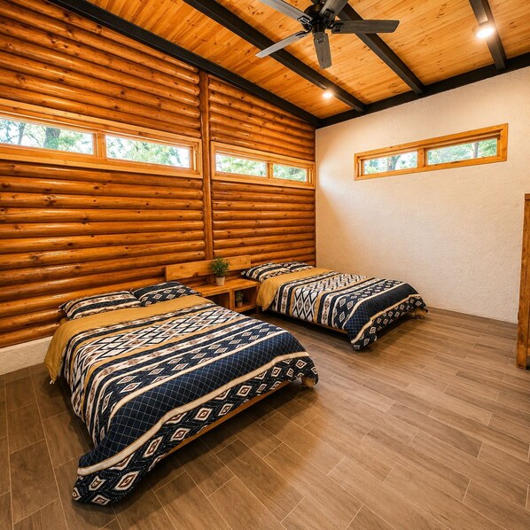 A log cabin bedroom with two beds, wooden walls, a ceiling fan, and windows.