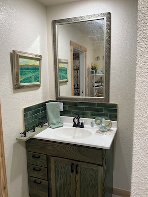 Une salle de bain avec un lavabo blanc, un mur recouvert de carreaux verts, un meuble en bois et deux tableaux encadrés.