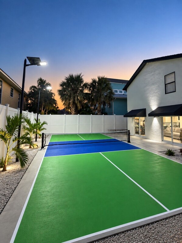 An outdoor tennis court with a net, surrounded by a white fence, palm trees, and a building with a balcony. - Siesta Key Beach House | Arcade, Pickleball, Pool (Sarasota)