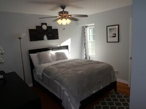 Un dormitorio con una cama, ventilador de techo, mesita de noche, lámpara y ventana con cortinas.