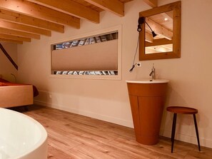 Ein modernes Badezimmer mit Holzboden, einer großzügigen Badewanne, einem hölzernen Waschtisch und einem Fenster mit Jalousien.