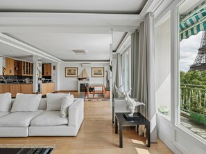 A modern living room with a white sofa, a dining area with a table and chairs, and a view of the Eiffel Tower through large windows. - Passy - Pdt. Kennedy 2 bd by B'Your Home (Paris)