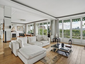 A modern living room with a sectional sofa, a dining area with a table and chairs, and large windows offering a city view. - Passy - Pdt. Kennedy 2 bd by B'Your Home (Paris)