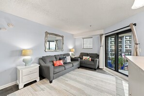 A modern living room with a grey sofa, a white side table, a mirror, and a view of the outside through a sliding glass door. - Sun and Splash Myrtle Beach Resort Condo (Myrtle Beach)