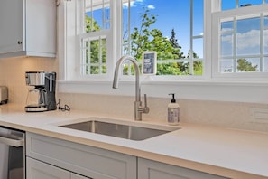 A kitchen with a stainless steel sink, a coffee maker, and a window with a view of greenery. - Blue Rocks Ocean Bliss for 10 (South Shore NS)