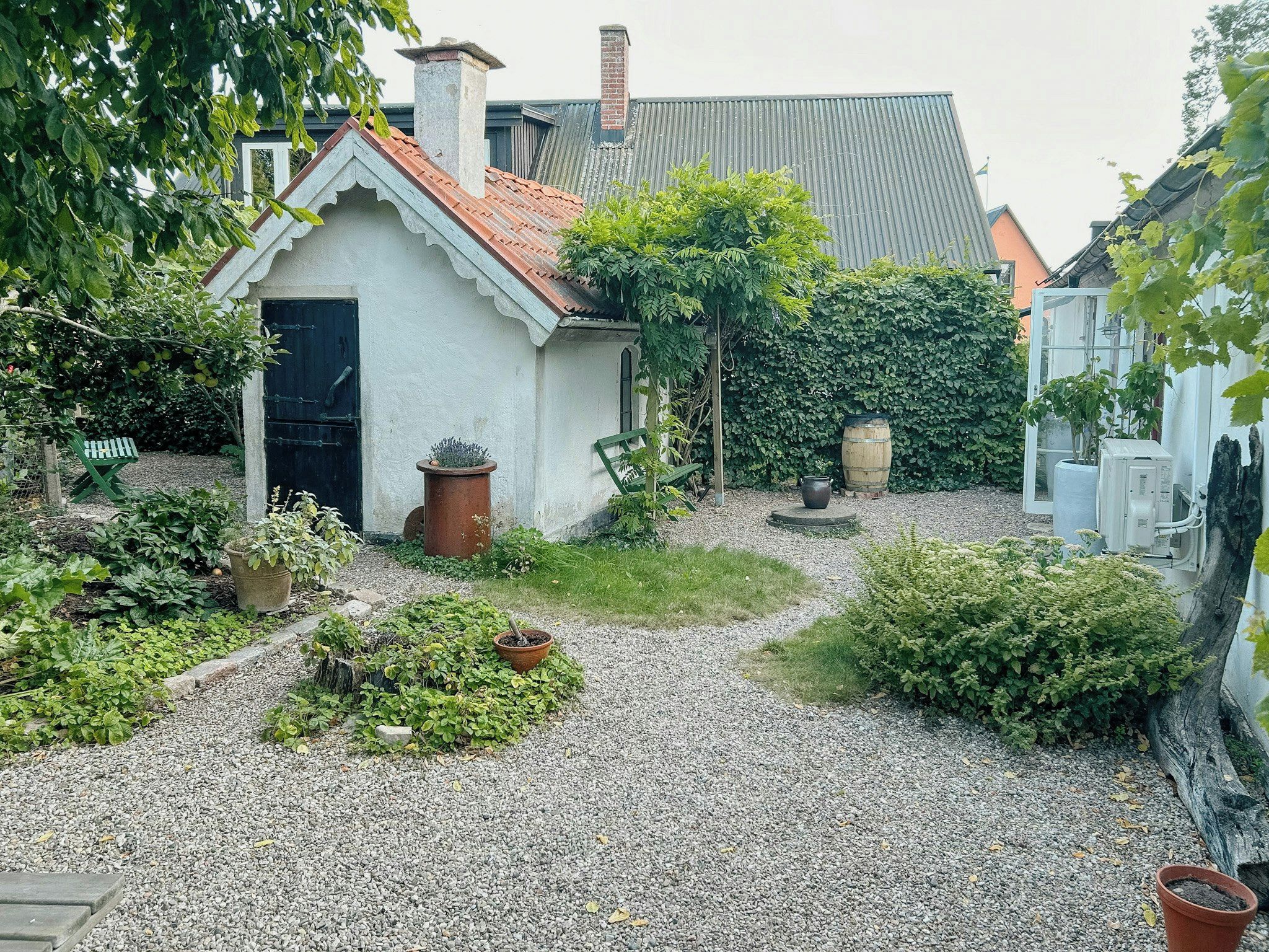 A small white building with a red-tiled roof, surrounded by gravel, plants, and a wooden barrel.