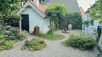A small white building with a red-tiled roof, surrounded by gravel, plants, and a wooden barrel.