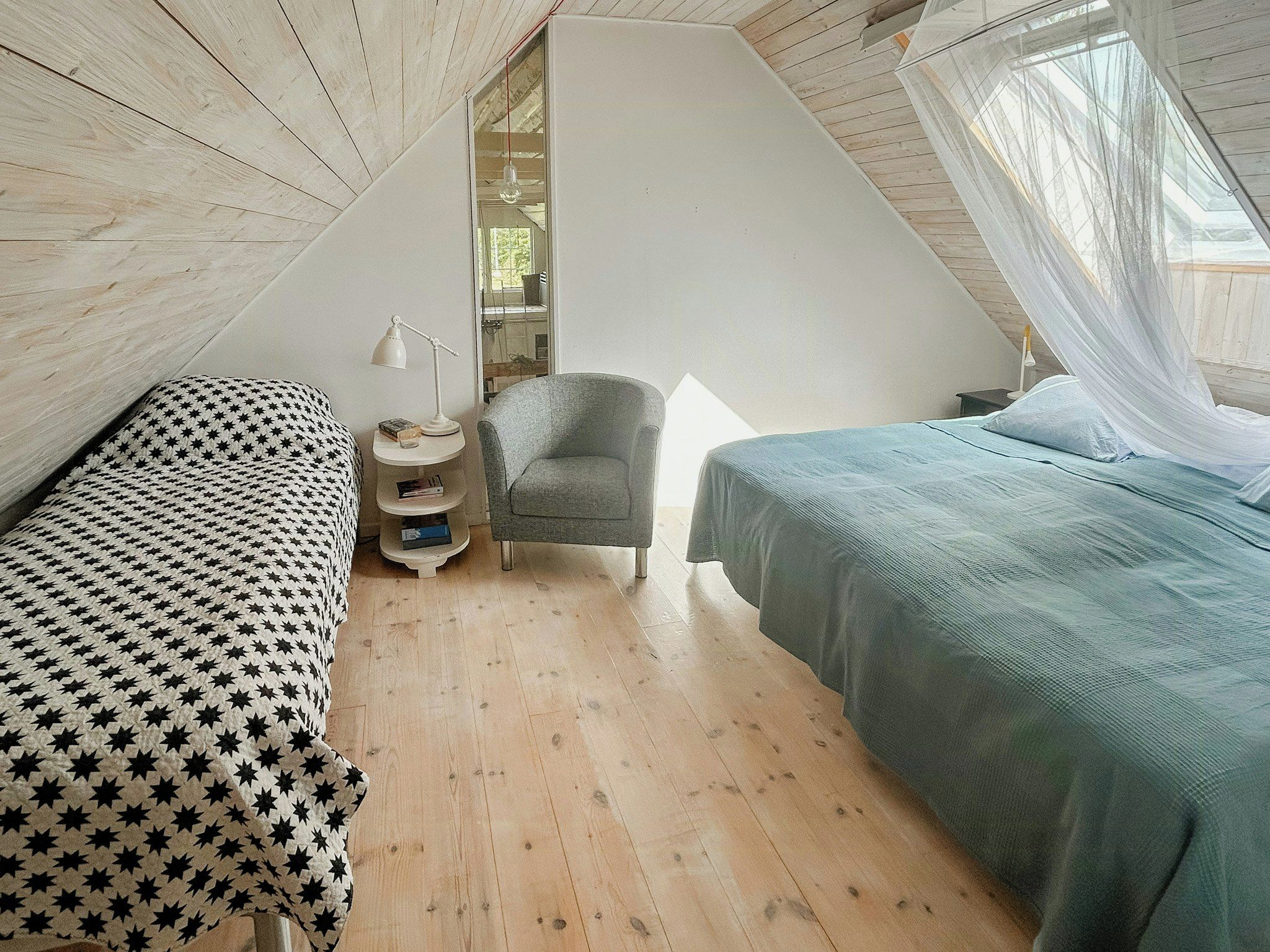 A bedroom with two beds, a chair, a bedside table with a lamp, and a window with a view of trees.
