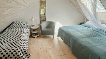 A bedroom with two beds, a chair, a bedside table with a lamp, and a window with a view of trees.