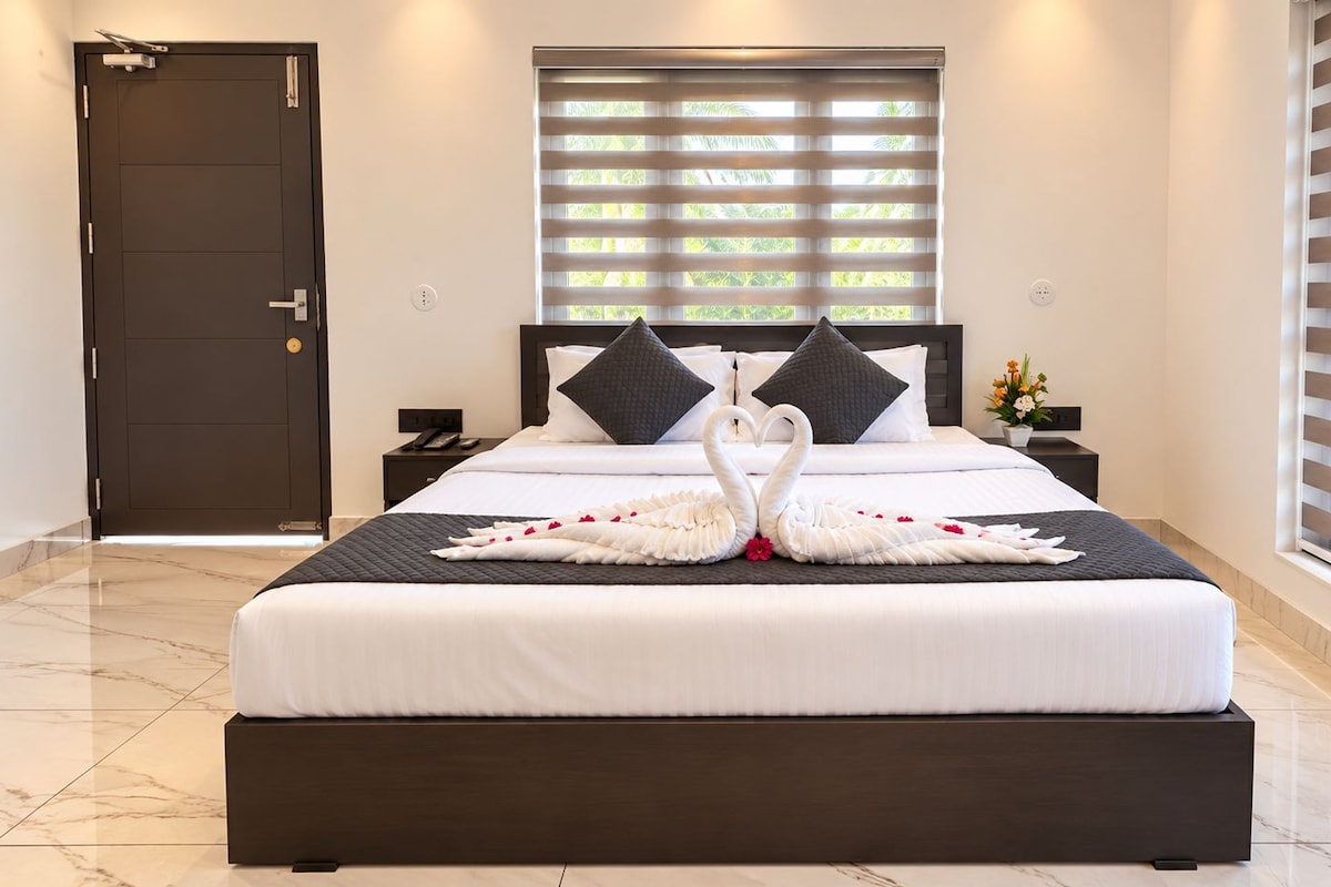 A hotel room with a large bed, a dark door, a window with blinds, and a nightstand with a vase of flowers.