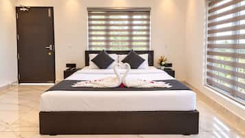 A hotel room with a large bed, a dark door, a window with blinds, and a nightstand with a vase of flowers.
