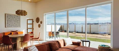 A modern living room with a large window, a sectional sofa, a dining area, and a ceiling fan.