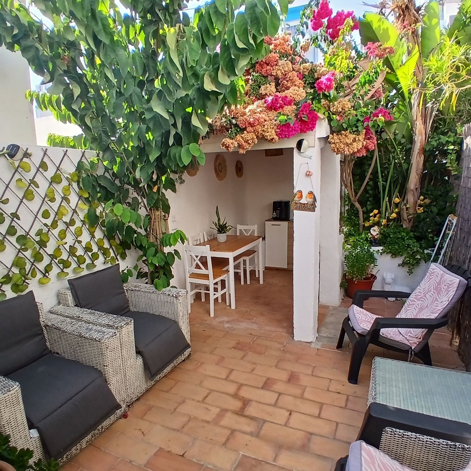 Un espacio de patio con una mesa y sillas, rodeado de plantas en macetas y un adorno de pájaro colgado en la pared.