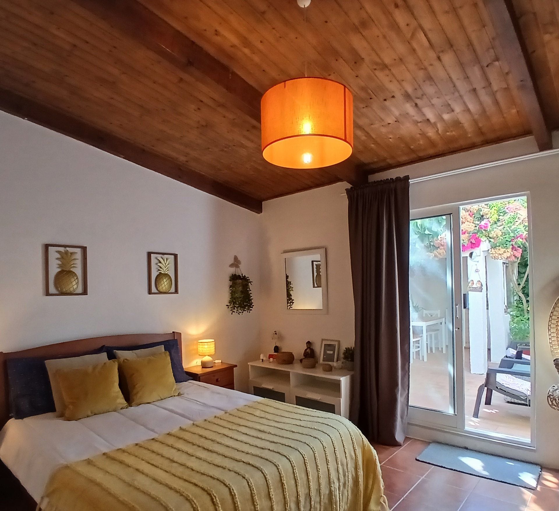 Un dormitorio con techo de madera, una cama con manta a rayas amarillas, una mesita de noche con lámpara, un espejo y vistas a un jardín a través de una puerta corrediza de vidrio.