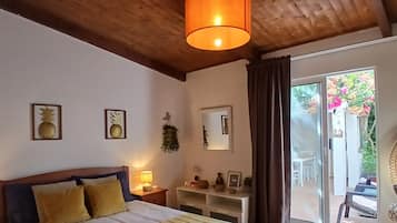 Un dormitorio con techo de madera, una cama con manta a rayas amarillas, una mesita de noche con lámpara, un espejo y vistas a un jardín a través de una puerta corrediza de vidrio.