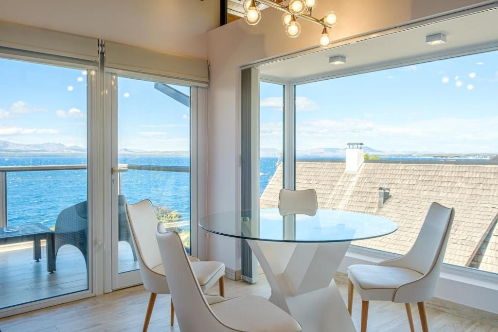 A modern room with a glass table and chairs, large windows with a view of the sea, and a cityscape in the distance.