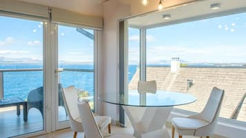 A modern room with a glass table and chairs, large windows with a view of the sea, and a cityscape in the distance.