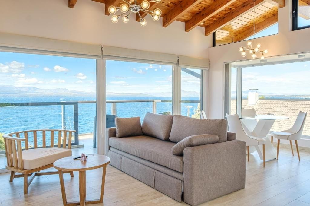 Ein modernes Wohnzimmer mit einer Couch, Sesseln und einem Tisch, großen Fenstern mit Meerblick und einer Holzbalkendecke.
