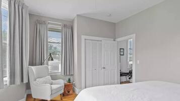 A bedroom with a large bed, a chair, a desk, and a window with curtains.