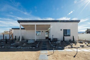 A modern house with a flat roof, a covered entryway, and a desert garden with rocks and cacti. - Midnight Sun | Custom pool, pickleball court, spa (Joshua Tree)