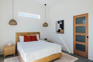 A bedroom with a wooden bed, a nightstand, a bedside table, a window, a door, and a wall-mounted artwork. - Midnight Sun | Custom pool, pickleball court, spa (Joshua Tree)