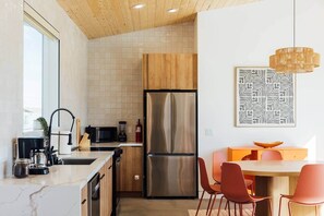 A modern kitchen with a stainless steel refrigerator, wooden cabinets, a microwave, and a dining area with orange chairs. - Midnight Sun | Custom pool, pickleball court, spa (Joshua Tree)