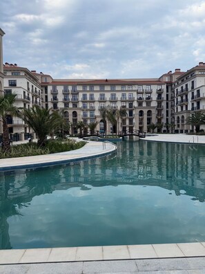 A swimming pool surrounded by multi-story buildings with balconies, palm trees, and a clear sky. - Marina Village: luxury 1-bedroom apartment by seaside in Nardaran, Baku (Nardaran)