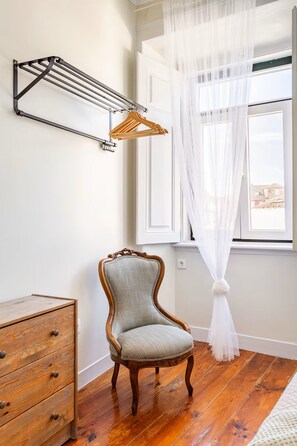 Una habitación con una silla de madera, una estantería metálica y una ventana con cortinas traslúcidas.
