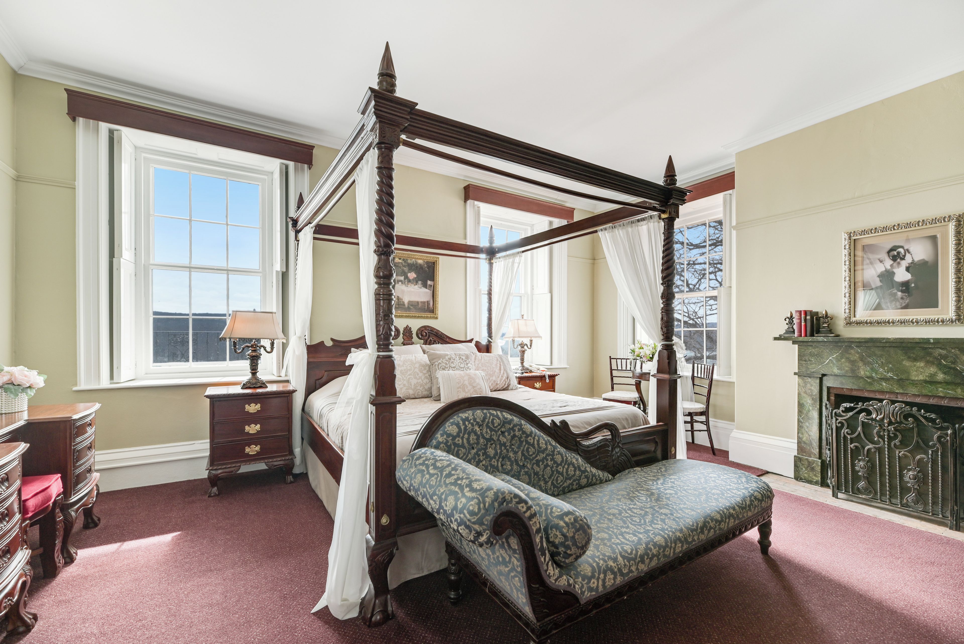 A four-poster bed with a canopy, a chair with a floral pattern, a fireplace, and a large window with a view.