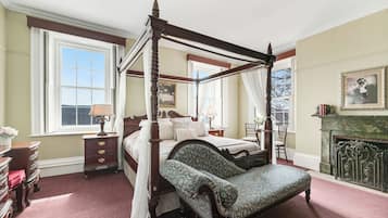 A four-poster bed with a canopy, a chair with a floral pattern, a fireplace, and a large window with a view.