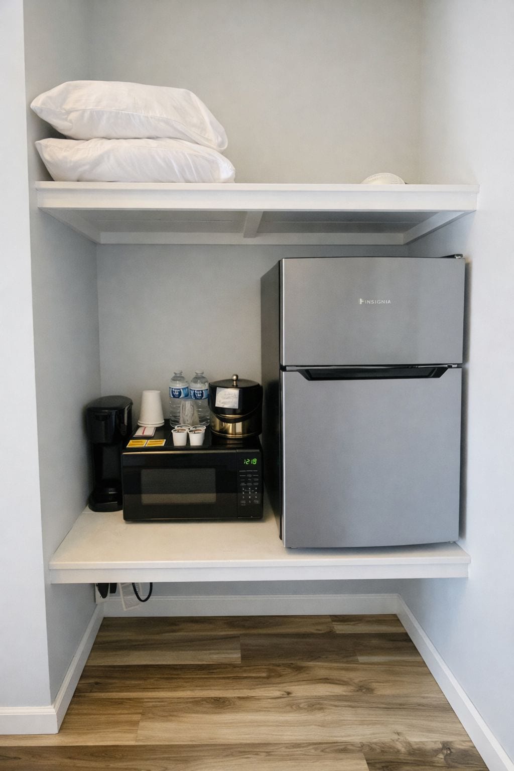 Une petite cuisine équipée d’un réfrigérateur en acier inoxydable, d’un four à micro-ondes noir et d’une machine à café posée sur une étagère blanche.