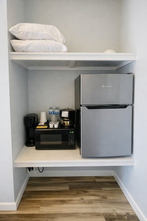 Una kitchenette con un refrigerador de acero inoxidable, un microondas negro y una cafetera sobre un estante blanco.
