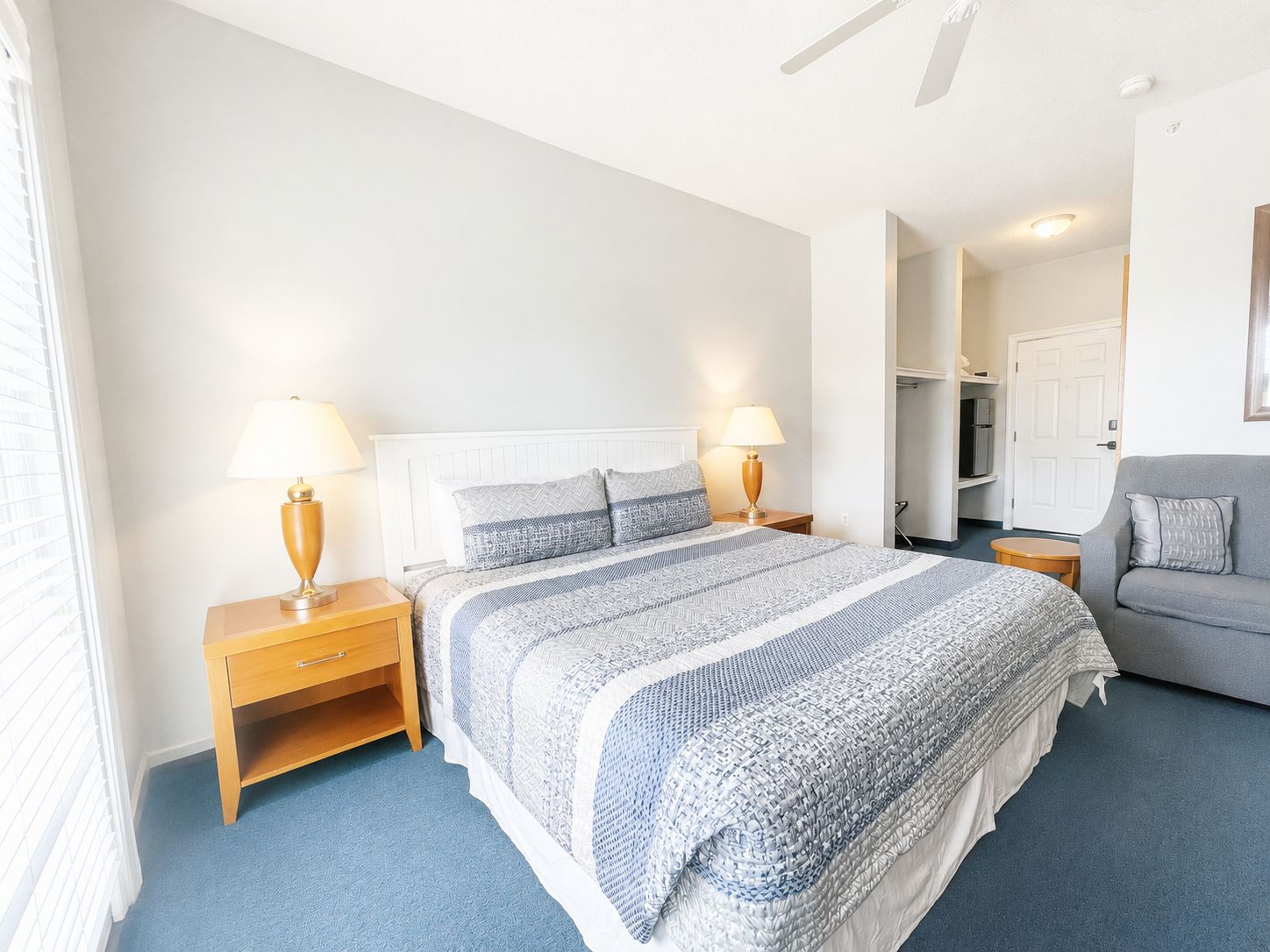 A bedroom with a bed, two bedside tables with lamps, a grey sofa, and a door leading to a bathroom.